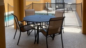 Solrig udendørs terrasse med et rundt bord og stole, en swimmingpool og udsigt over et boligområde.