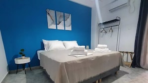 Ein ordentlich bezogenes Bett mit weißen Leinen, ein kleiner Nachttisch mit Pflanze und eine blaue Wand mit abstrakter Kunst.