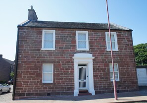 A two-story brick building with a white front door, two windows on the upper floor, and a garage door on the right. - Fantastic 2 Bedroom, Spacious and Great Location! (Helensburgh)