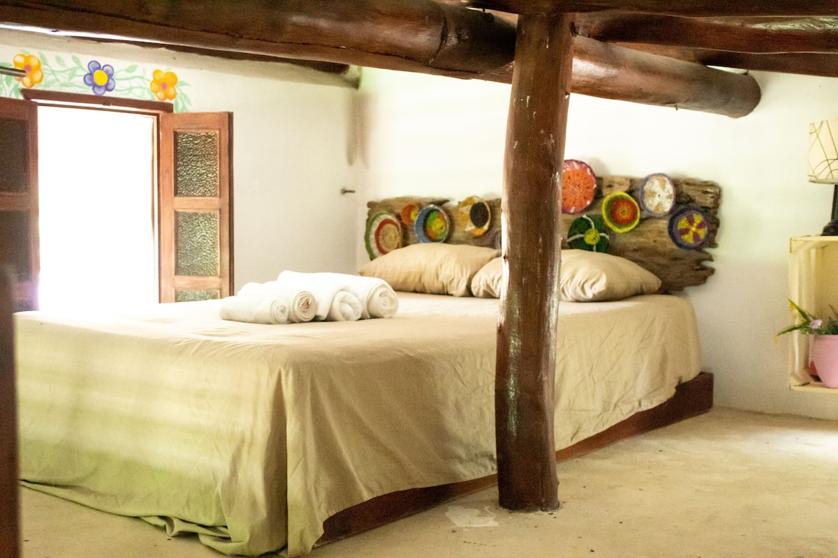 Un lit avec une couvre-lit beige, des oreillers et des serviettes enroulées. Une poutre en bois se trouve au-dessus du lit.