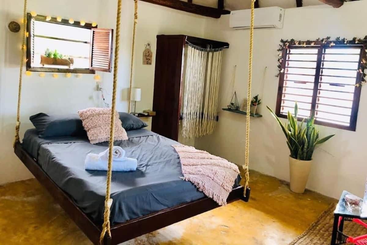Une pièce avec un lit suspendu au plafond par des cordes, une fenêtre avec des stores, une plante en pot et une petite table avec une chaise.