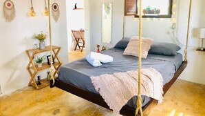 A bedroom with a hanging bed, a wooden floor, a small table with plants, and a chair. - Cabañas Manati (Isla Holbox)