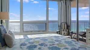 Een slaapkamer met een groot raam met uitzicht op de oceaan, een bed met een bloemenbedekking, een stoel en een nachtkastje.