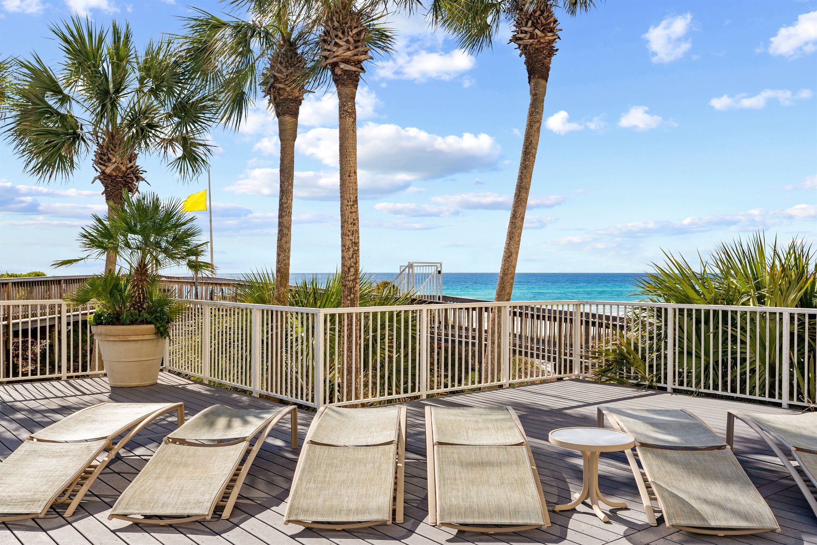 Eine Terrasse mit Liegestühlen und einem Blumentopf mit Blick auf einen Strand mit Palmen und einem klaren Himmel.