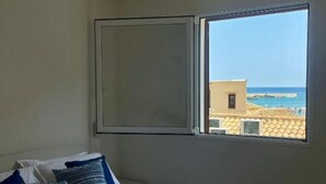 A bedroom with a bed, pillows, and a window with a view of buildings and a body of water. - Gassa d’Amante (Castellammare del Golfo)