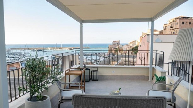 A balcony with wicker furniture, a potted plant, and a view of the sea and buildings. - Gassa d’Amante (Castellammare del Golfo)