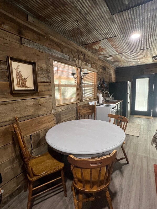 A rustic dining area with a round table and wooden chairs, a framed picture on the wall, and a kitchen in the background.