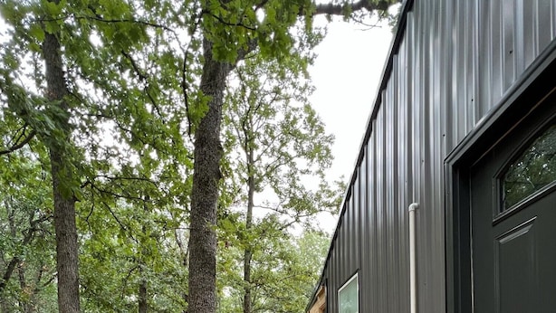 Une terrasse en bois avec des meubles en osier, un bâtiment en métal avec une porte sombre, et un paysage boisé en arrière-plan.