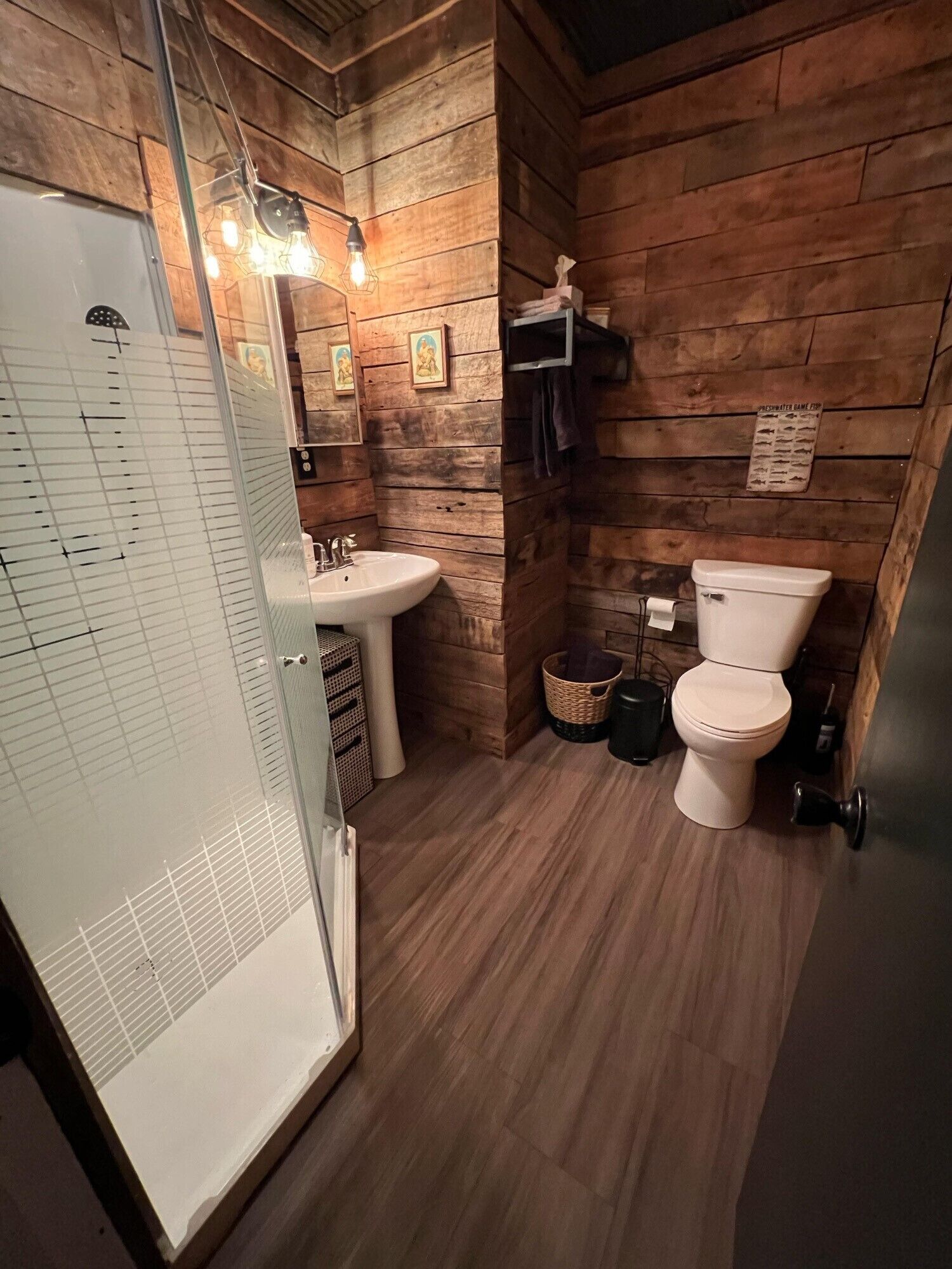 A compact bathroom with wooden walls, a white sink, a toilet, and a glass shower enclosure.