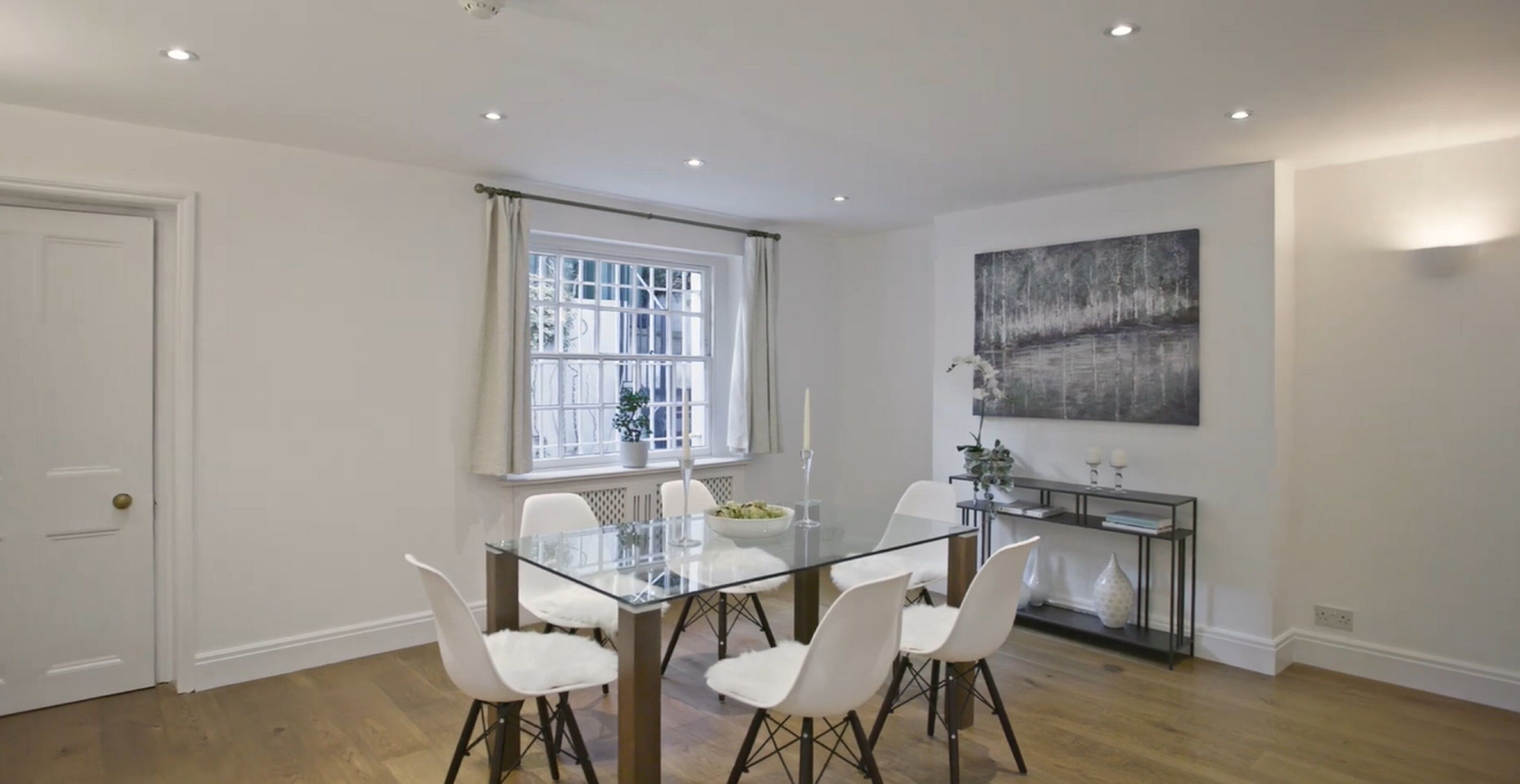 Un coin repas avec une table en verre et des chaises blanches, une fenêtre avec des rideaux, un tableau au mur et une petite plante posée sur la table d’appoint.