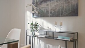 Ett modernt rum med ett glasbord, en vit stol, ett avlastningsbord med böcker och ljus samt en stor, abstrakt målning på väggen.