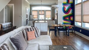 A spacious living room with a large sofa, a dining area with a wooden table and chairs, a kitchen with modern appliances, and a colorful abstract wall. - MusikHouse East I Rooftop + Hot Tub I 5 mi to DWNT (Nashville)