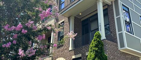 Un edificio de ladrillo de varios pisos con un balcón, un césped bien cuidado y árboles floreados.