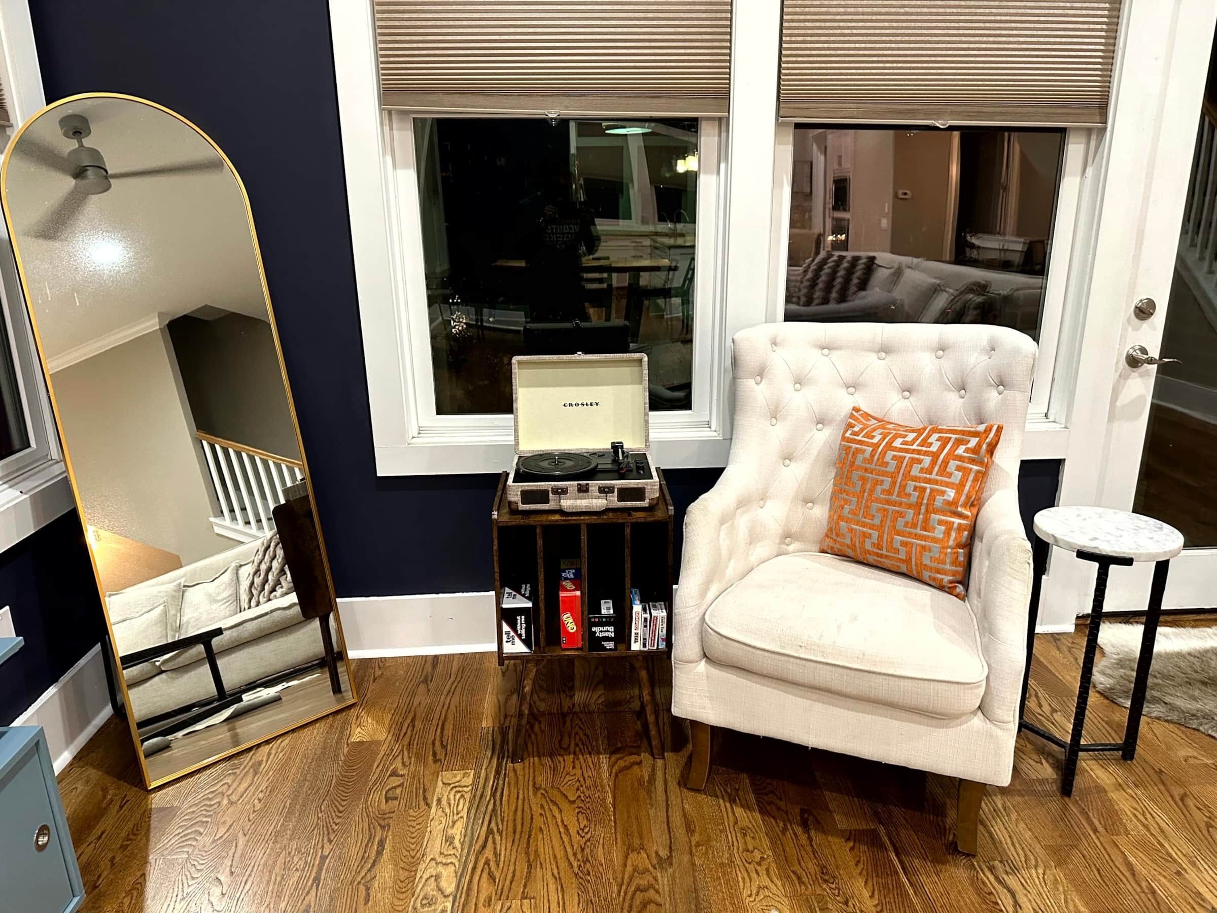A room with a mirrored wardrobe, a white armchair with a patterned cushion, a small table with a laptop, and a wooden floor.