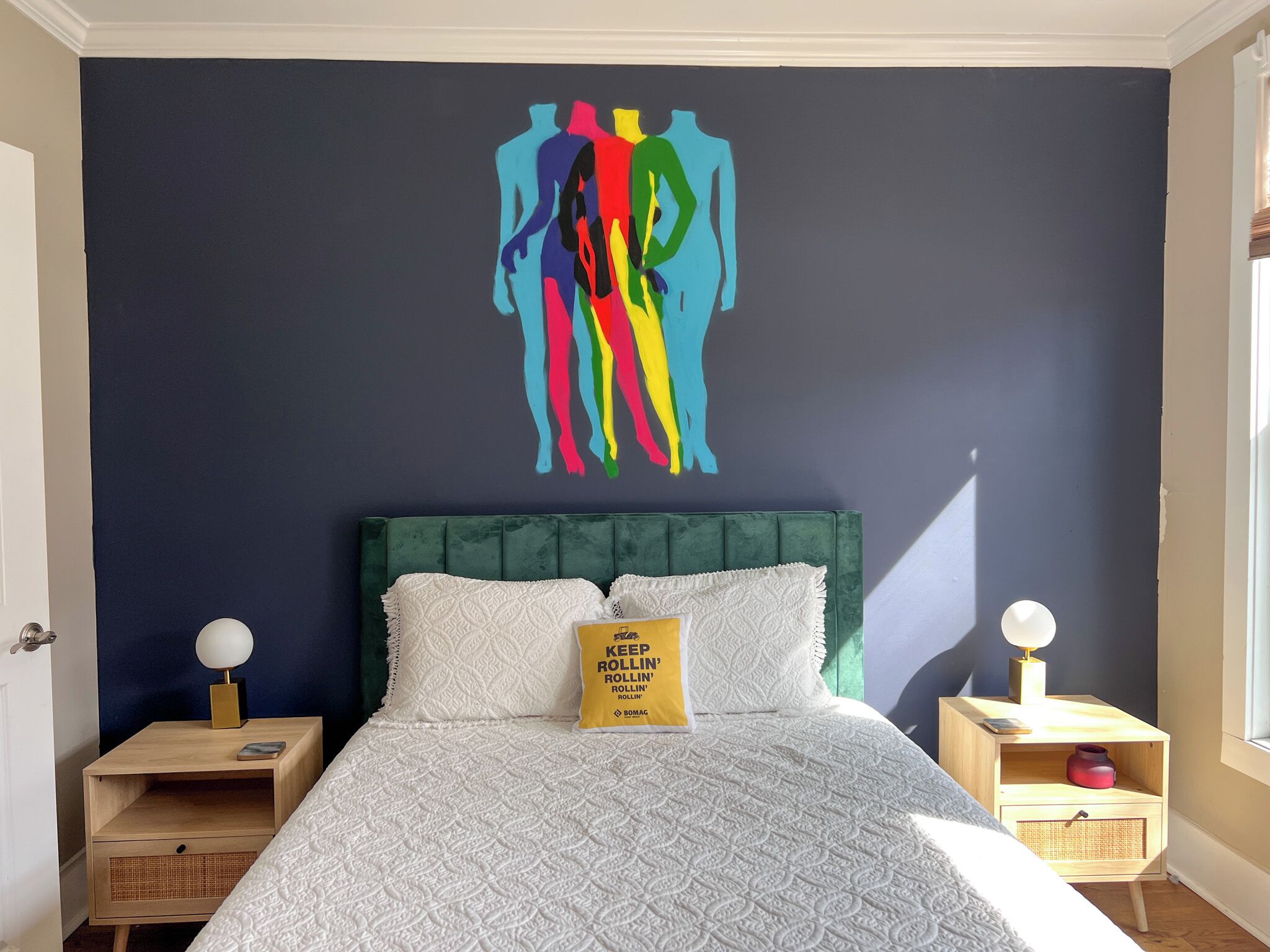 A bedroom with a dark blue wall, a green upholstered headboard, white textured bedding, and a yellow pillow with text.