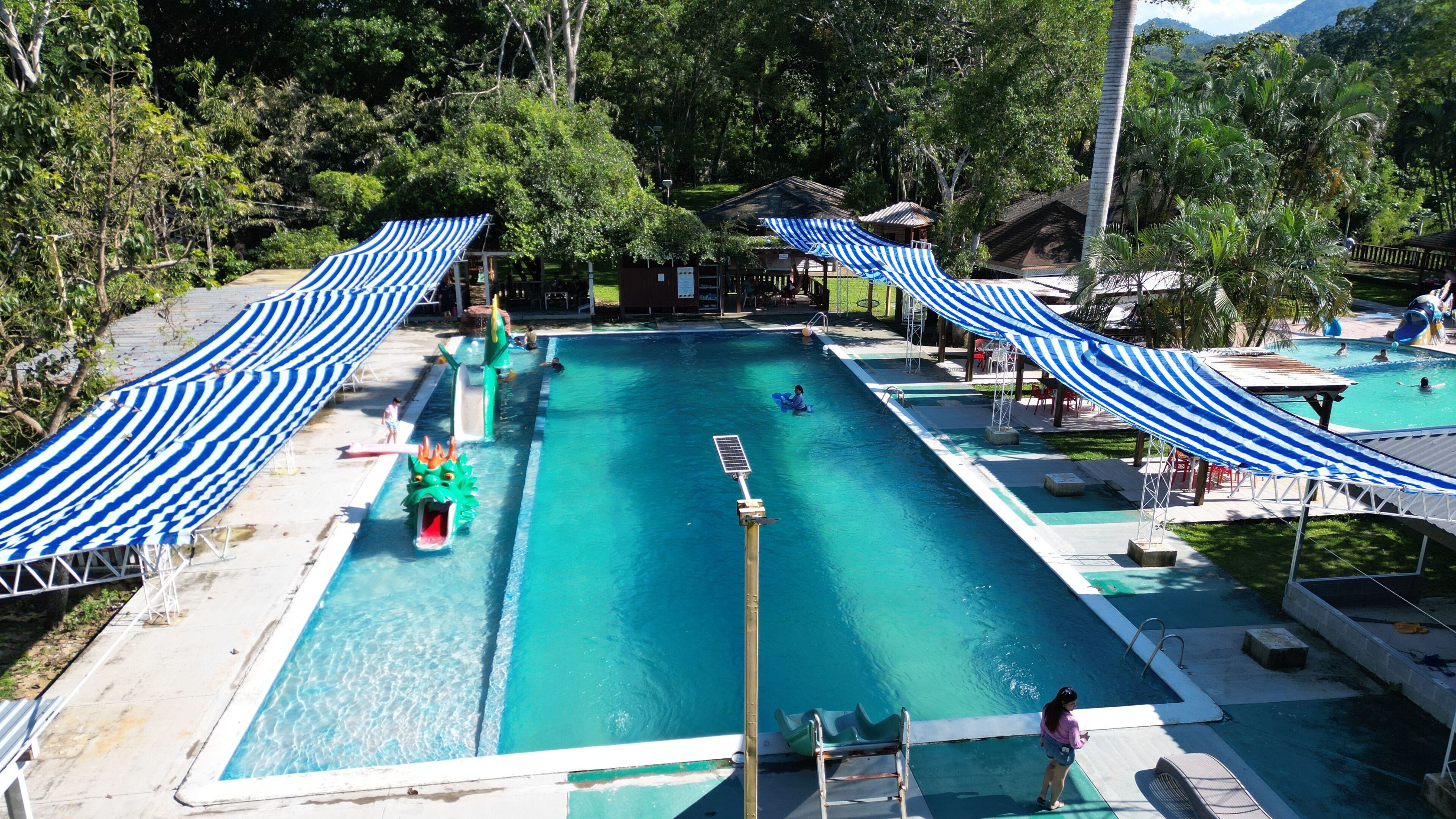 Una piscina con tobogán, rodeada de árboles y cubierta por toldos a rayas.