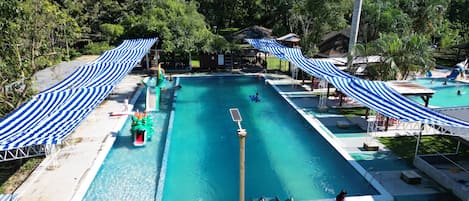 A swimming pool with a slide, surrounded by trees and covered by striped canopies.