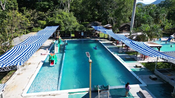 A swimming pool with a slide, surrounded by trees and covered by striped canopies. - Hotel Cabañas River Park (Omoa)