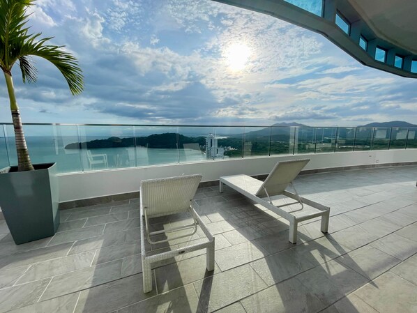 A balcony with two white wicker chairs, a potted plant, and a glass railing overlooking a scenic view.