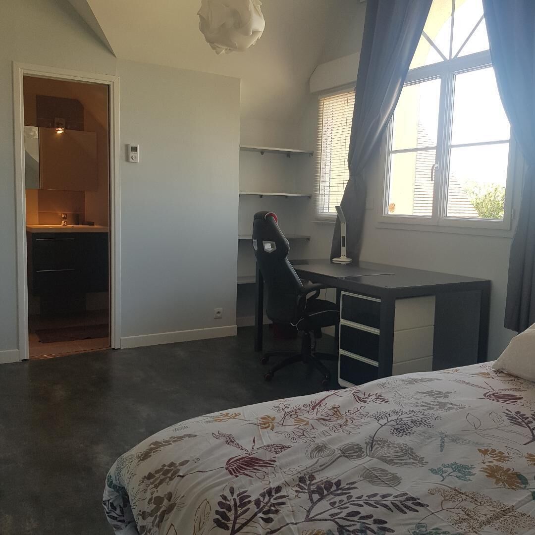 A bedroom with a bed, a desk, and a chair. There is a window with curtains, a door leading to a bathroom, and a ceiling light fixture.