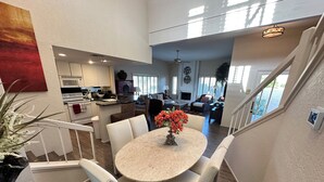 Una cocina moderna con armarios blancos, un comedor con una mesa de mármol y sillas blancas, y una escalera que lleva a una sala de estar con chimenea.