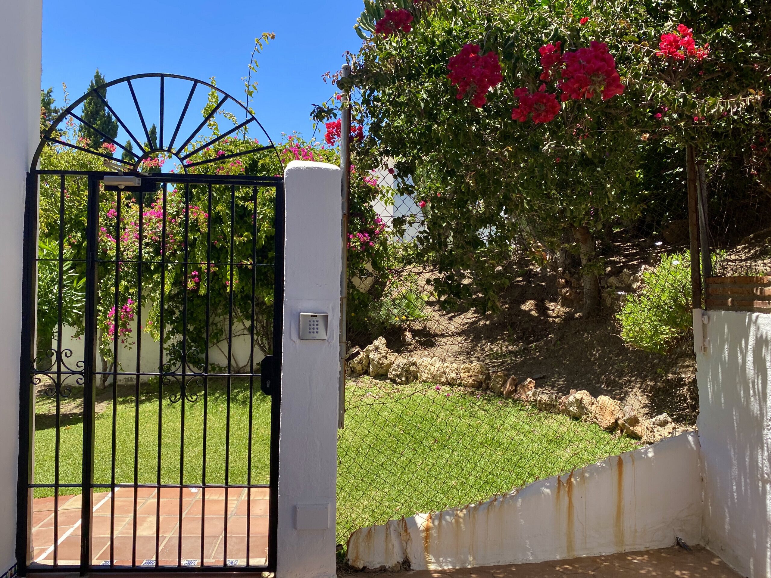 A gated entrance with a metal gate and a keypad lock, surrounded by a white wall and lush greenery.
