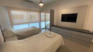 Habitación de hotel con dos camas, un televisor montado en la pared y un ventanal grande con persianas.