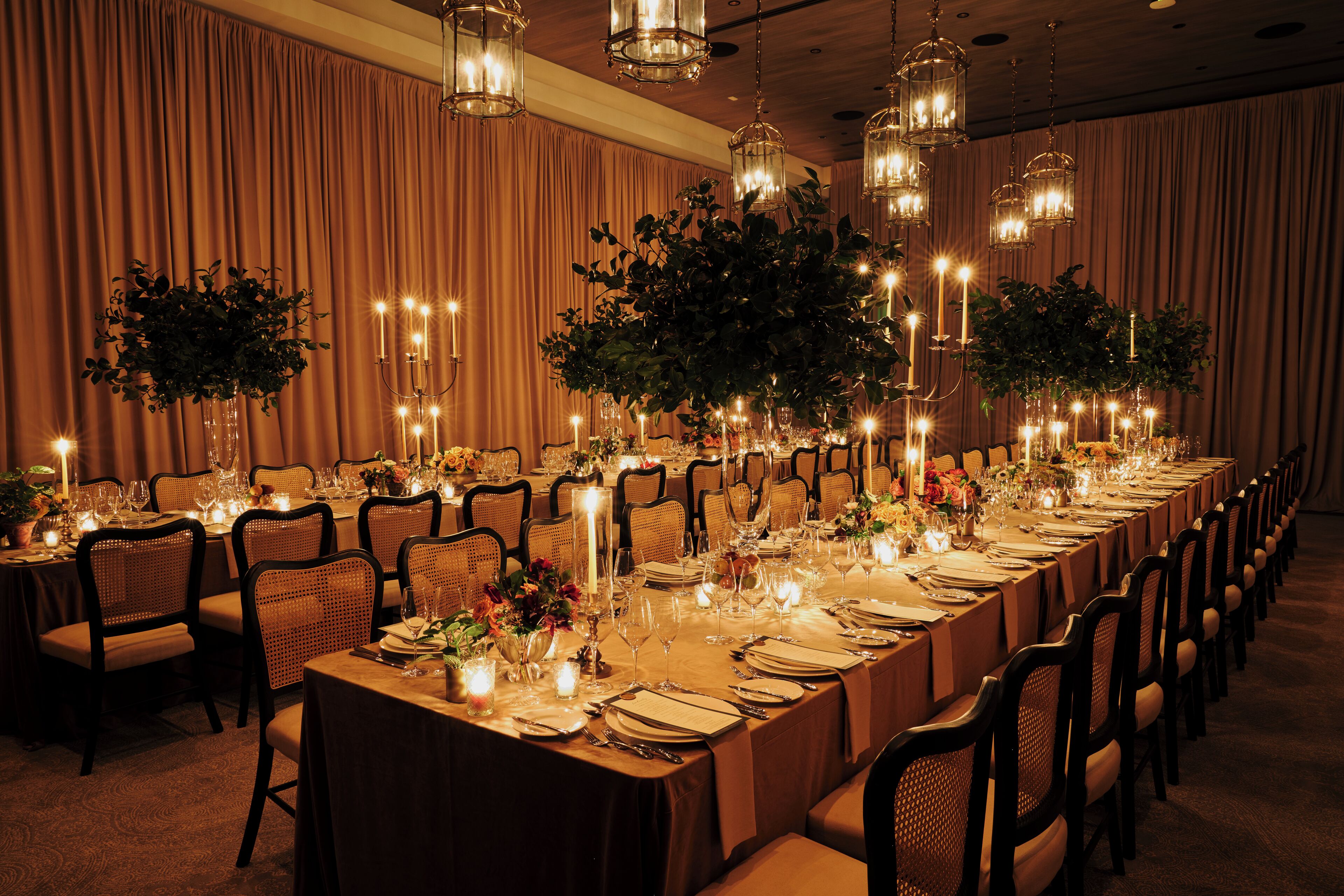 A long banquet table set for a formal event, with chairs on both sides, candles, and floral centerpieces.