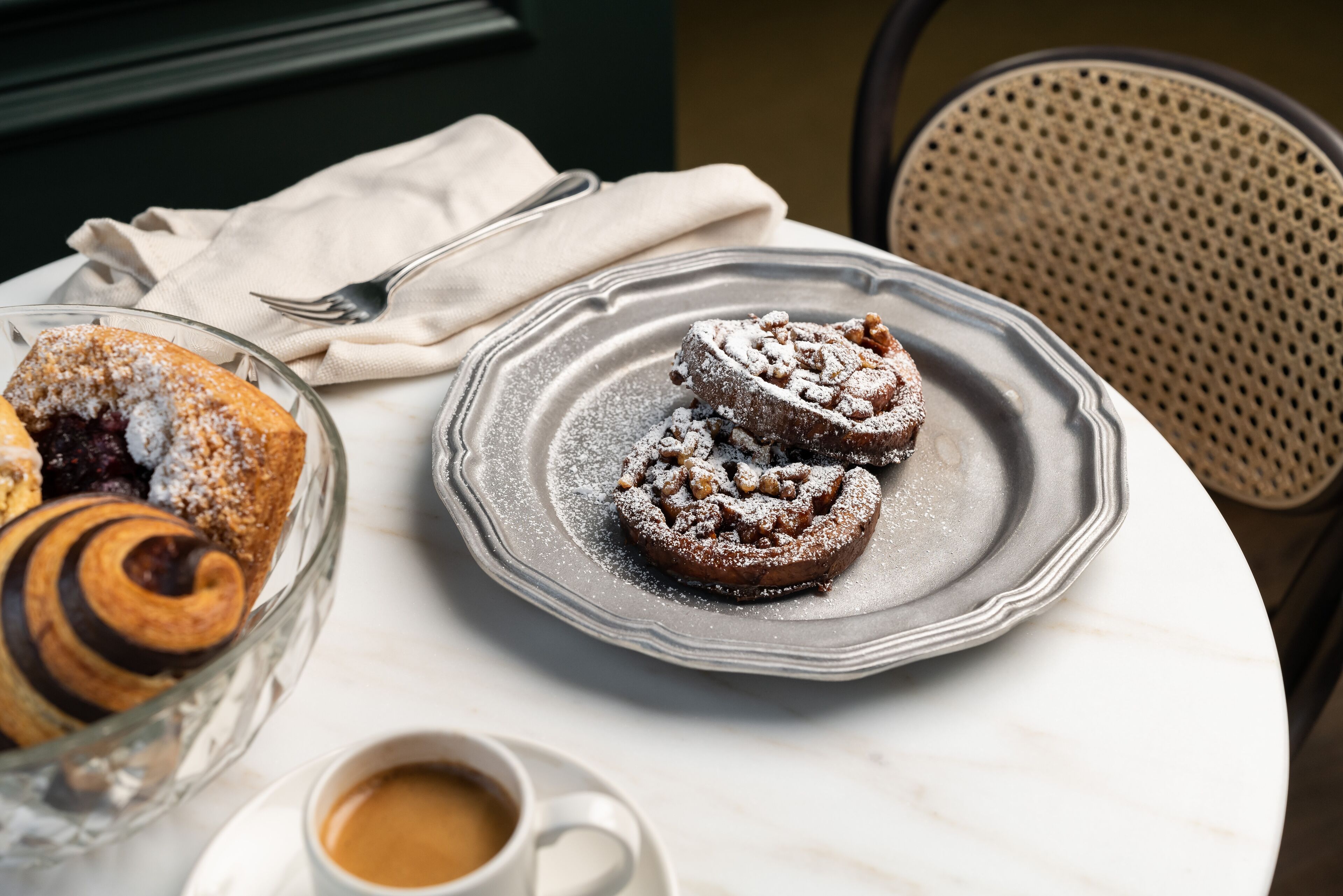 Una mesa con un plato de rollos de canela y un bol de pasteles, una taza de café y un tenedor sobre una servilleta.