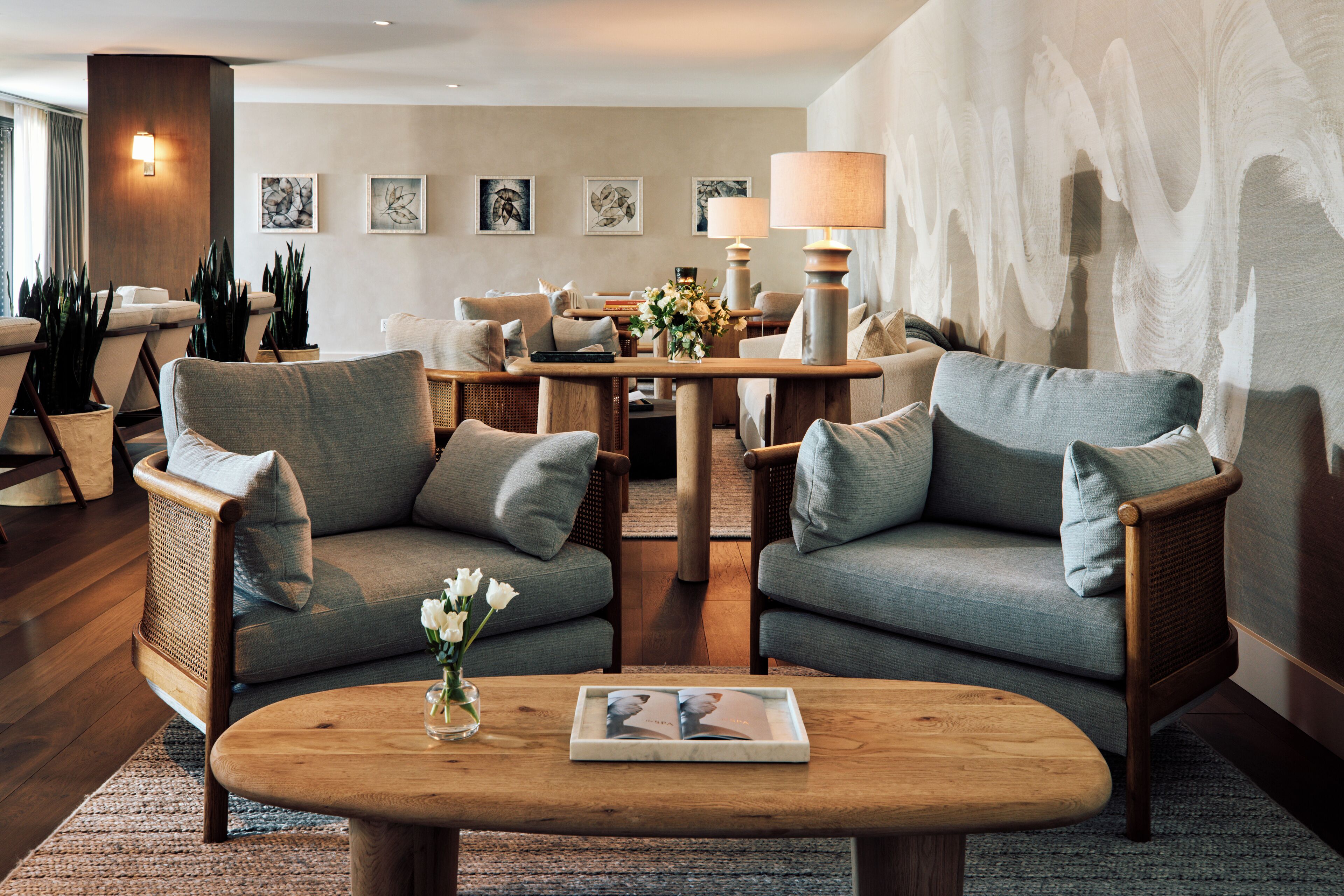 A modern living room with two sofas, a coffee table, and a dining table with lamps. There are potted plants and artwork on the walls.