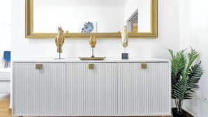 A white, ribbed console with gold handles, a gold-framed mirror above it, and decorative items including a golden bird and a feather.