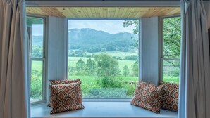 Ein Zimmer mit großem Fenster mit Blick auf eine grüne Landschaft und Hügel. Auf einer Bank liegen zwei Kissen mit Muster.