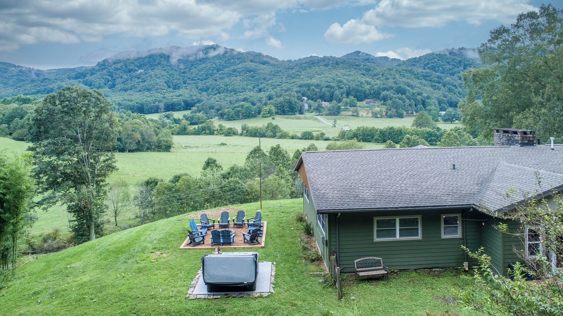 A green house with a sloped roof, a deck with outdoor furniture, and a view of a green valley and mountains in the background.