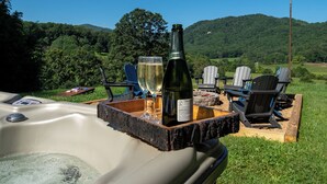 Ein Whirlpool mit einer Holztischplatte, auf der eine Weinflasche und zwei Gläser stehen, umgeben von Adirondack-Stühlen in einer grünen Wiese mit einem bewaldeten Hügel im Hintergrund.