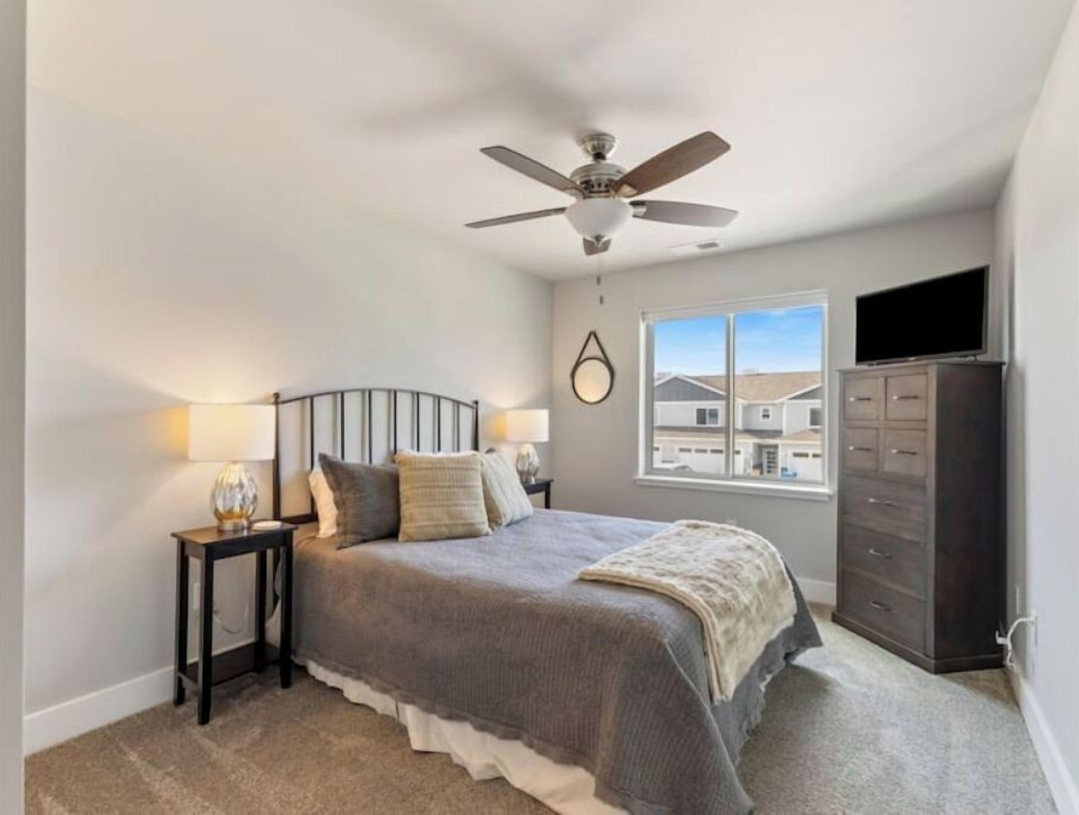 A bedroom with a bed, a nightstand, a dresser, a ceiling fan, and a window with a view of houses.