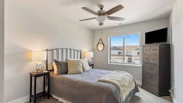 A bedroom with a bed, a nightstand, a dresser, a ceiling fan, and a window with a view of houses.