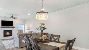 A modern living room with a fireplace, a dining area with a chandelier, and a ceiling fan. - Townhome Near Bozeman Airport (Belgrade)
