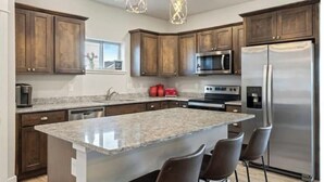 A modern kitchen with dark wooden cabinets, stainless steel appliances, and a granite island with bar stools. - Townhome Near Bozeman Airport (Belgrade)