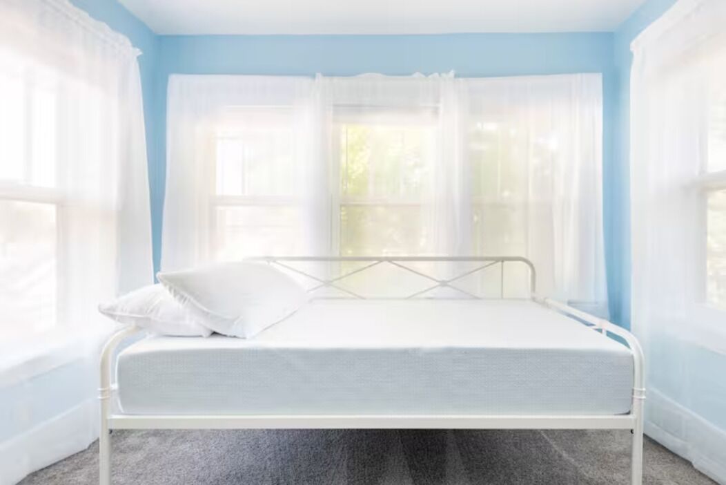 Uma cama de solteiro num quarto com paredes azul claro e cortinas brancas.