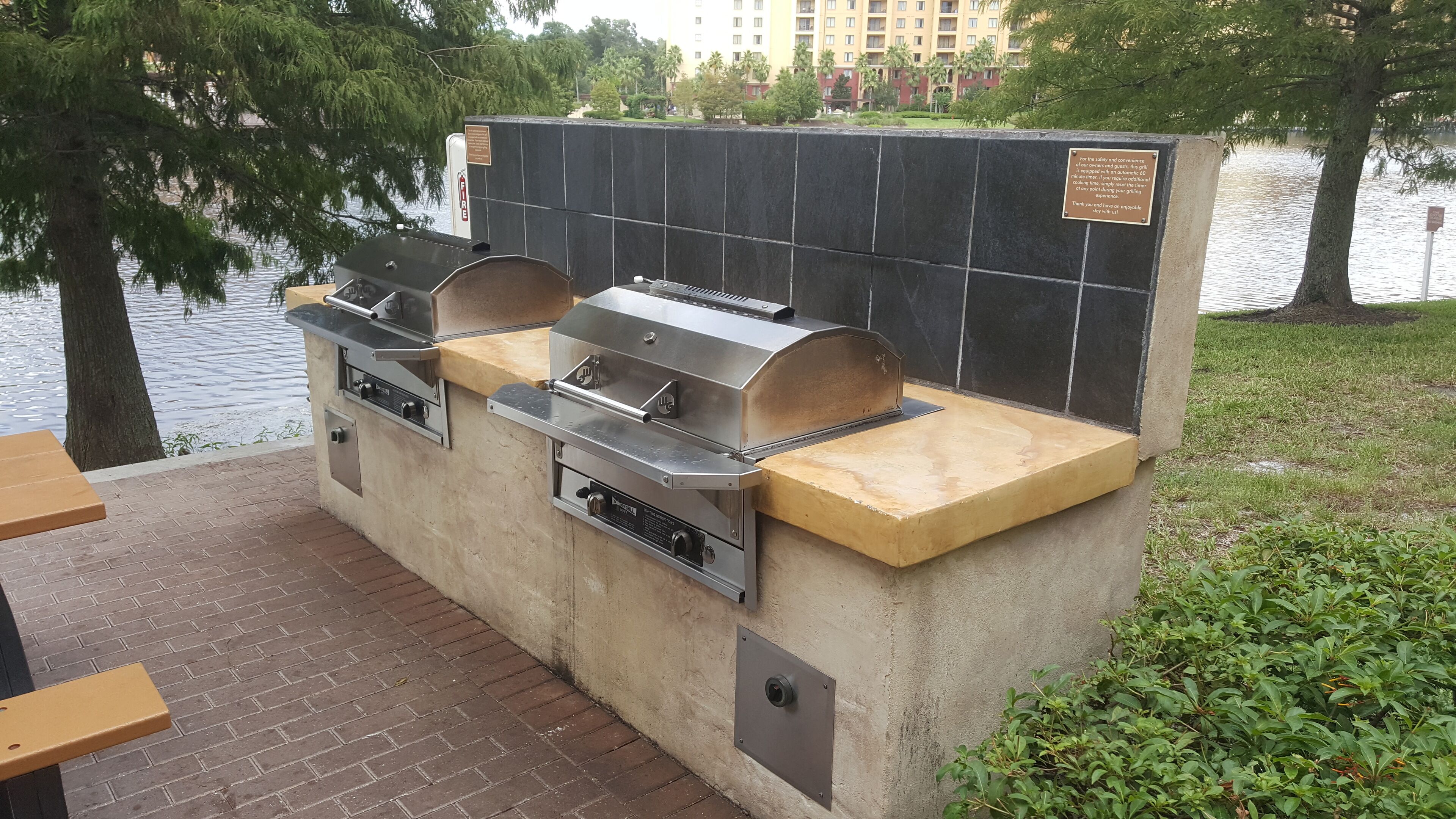 En udendørs grillplads med to gasgrill og en indbygget ovn, beliggende i nærheden af vand med udsigt til en bygning i baggrunden.