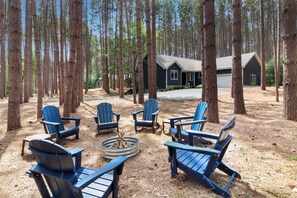 Een bebost gebied met verschillende Adirondack stoelen rond een vuurkuil. Een chalet is op de achtergrond zichtbaar.