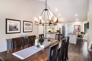 A spacious dining area with a wooden table, chairs, and a chandelier. There are framed pictures on the wall and a kitchen in the background.
