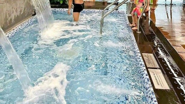 Ein Schwimmbad mit Wasserfall, umgeben von einer gefliesten Terrasse und Innenwänden.