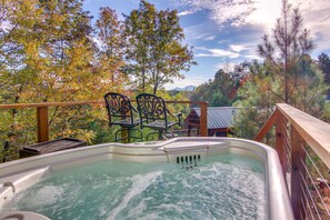 Un jacuzzi en una terraza de madera con sillas, rodeado de árboles y vistas a la montaña.