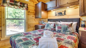 Une chambre à coucher dans un chalet en bois, avec un grand lit, une fenêtre avec des rideaux et une tête de lit ornée d’un motif représentant un ours et un arbre.