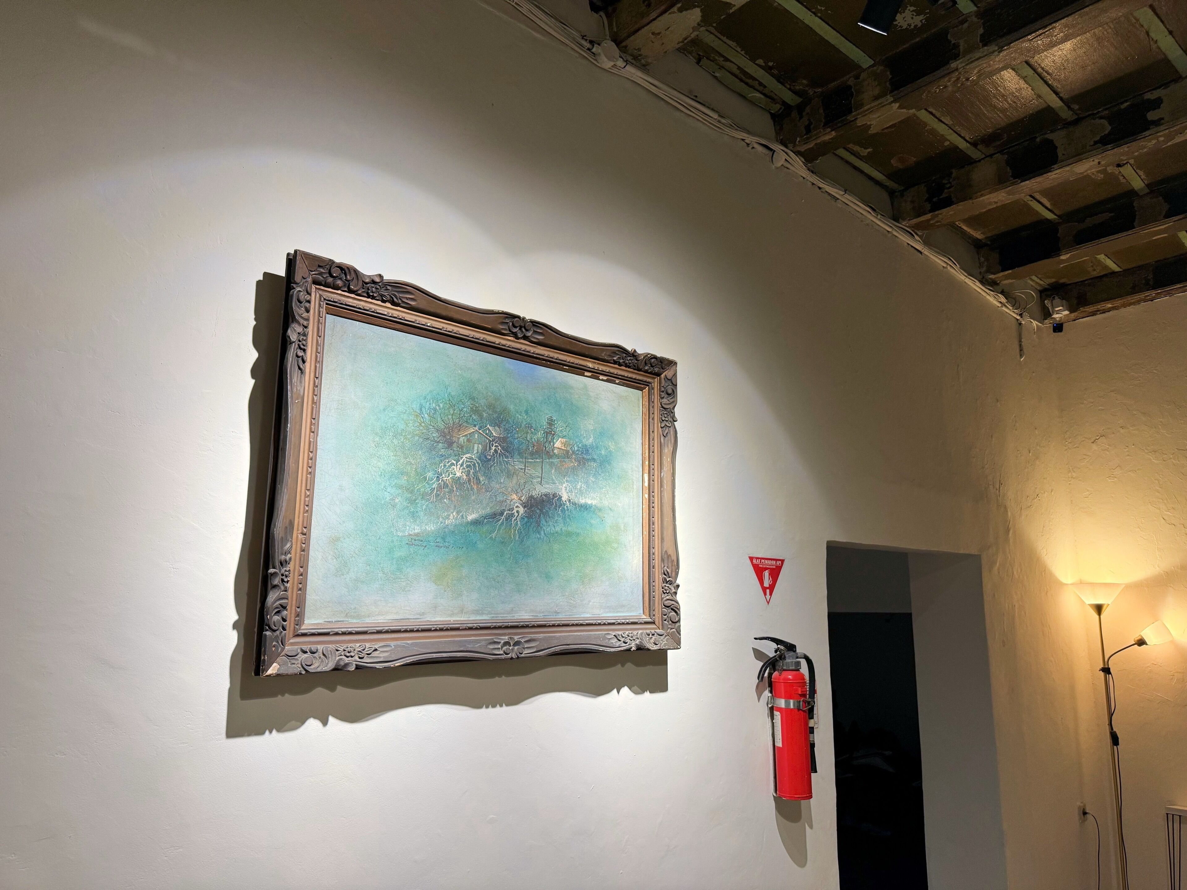A framed painting on a wall, a fire extinguisher, and a lit floor lamp.