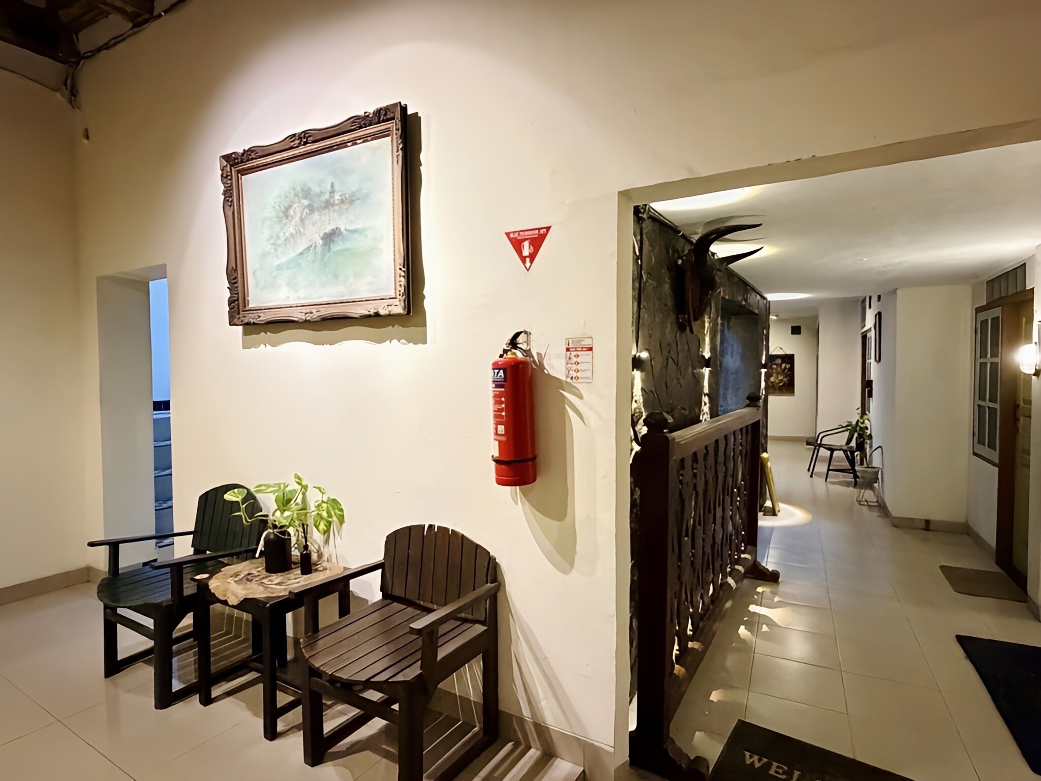 Un couloir avec un tableau encadré, un extincteur, une table avec des chaises et une porte sur laquelle est posé un paillasson avec l’inscription « Bienvenue ».