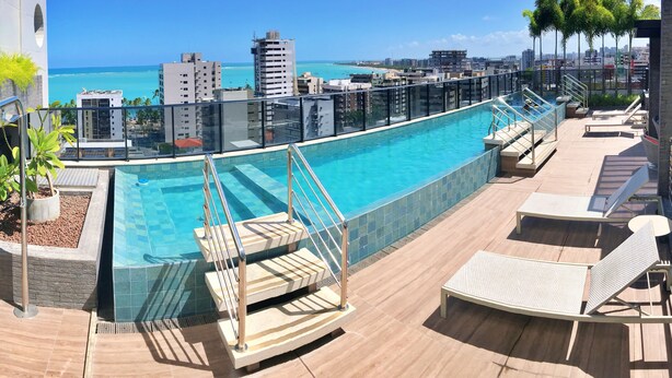 Une piscine sur le toit, entourée d’une paroi en verre, avec des chaises longues et une vue sur l’océan et le panorama urbain.