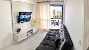 Un salon moderne avec un téléviseur à écran plat fixé à un mur en bois, un canapé gris, une petite plante et une vue sur le balcon extérieur.