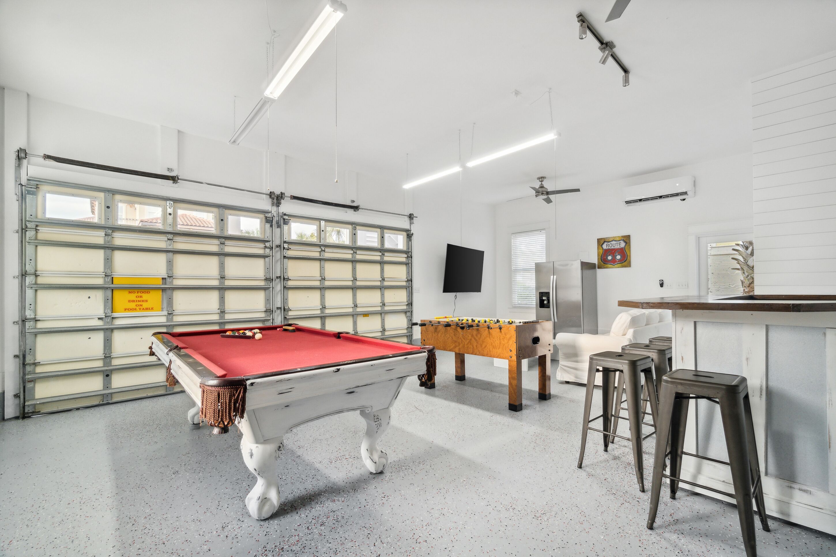 A pool table, foosball table, and bar area in a well-lit indoor space with a garage door and white walls.